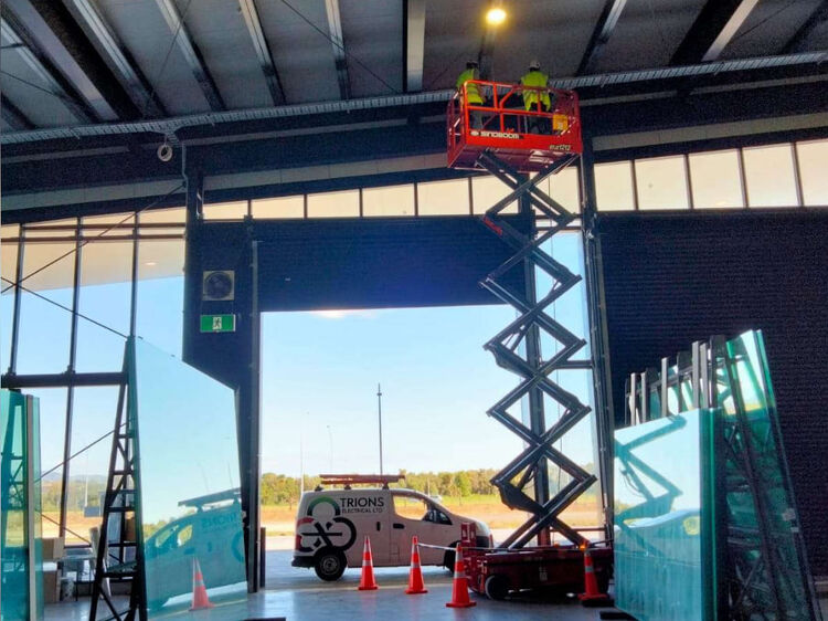 Electricians on a raised scissor lift installing overhead lighting inside a warehouse, with Trions van visible outside.