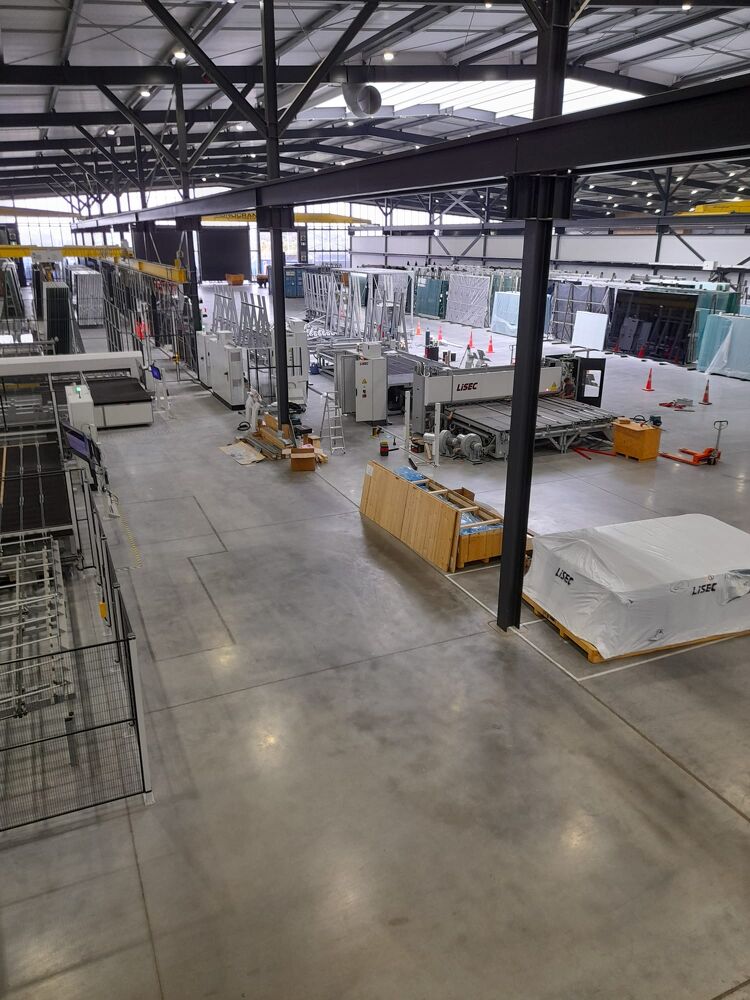 Wide view of a large industrial warehouse showing machinery, stacked glass panels, and electrical fit-out in progress.