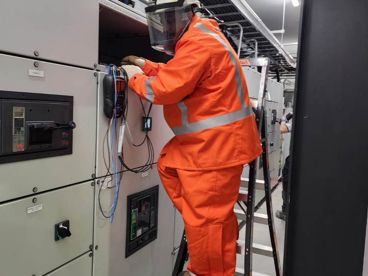 Electrician in full PPE and face shield working on high-voltage equipment from a ladder inside a control room.