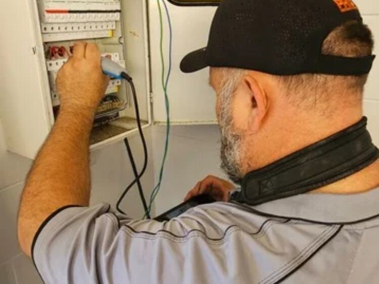 Trions Electrical technician testing wiring inside a wall-mounted switchboard using handheld equipment.